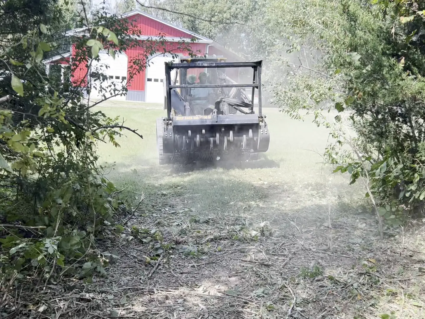 Forestry mulcher clearing brush along a lane with a red barn behind it
